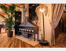 A cozy indoor space featuring a metallic stove with pots arranged in a row underneath, set against a partially charred brick wall. The area is adorned with a potted palm plant, a palm-shaped decorative floor lamp, and patterned curtains, with wooden flooring completing the warm ambiance.