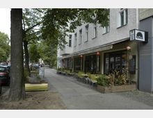 Gehweg mit einem Café, dessen Außenbereich mit grünen Stühlen und Tischen bestückt ist, vor einer Häuserfassade mit großen Fenstern und einem Schild. Links stehen Bäume und geparkte Autos entlang der Straße.