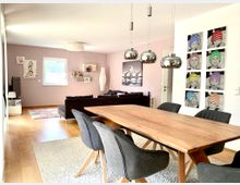 Ein modern eingerichtetes Wohnzimmer mit Essbereich. Im Vordergrund steht ein großer Holztisch mit grauen Polsterstühlen, darüber hängen drei silberne Hängeleuchten. Im Hintergrund befindet sich eine gemütliche Sitzecke mit schwarzem Sofa vor einer rosa Wand, dekoriert mit Fotos und einem Regal.