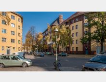 Straßenecke mit mehrstöckigen Wohngebäuden in gelber und rötlich-brauner Backsteinfassade; erkennbar sind geparkte Autos, ein Straßenschild mit der Aufschrift „Brahestraße“, ein Fahrrad an einer Laterne und Bäume mit herbstlichem Laub.