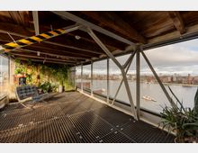 Industrieartig gestalteter Innenraum mit Gitterrostboden, sichtbaren Stahlträgern und Holzbalkendecke sowie einer begrünten Pflanzenwand. Große Fensterfront mit Blick auf einen breiten Fluss und gegenüberliegende Stadtbebauung.