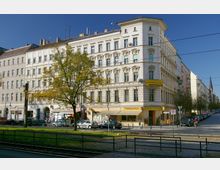 Historisches Wohn- und Geschäftsgebäude in einer städtischen Umgebung, mit einer hellen Fassade und dekorativen Fensterrahmungen. Im Erdgeschoss befinden sich Geschäfte und Restaurants, während im Vordergrund ein Baum, parkende Autos und Straßenbahnschienen zu sehen sind.