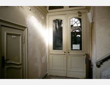 Ein abgenutzter Eingangsbereich mit einer weißen Doppeltür aus Holz, die Fenster mit bogenförmigen Rahmen enthält. Die Wände sind mit abgeblätterter Farbe bedeckt, daneben befindet sich eine schlichte Innentür mit dem Schild „Herren“.