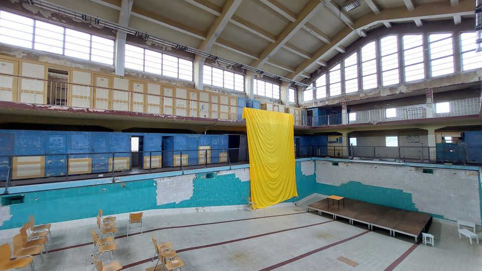 Ein leerstehendes Hallenbad mit hoher Decke und vielen Fenstern. Das Becken ist trocken, an den Wänden sind blaue und grüne Fliesen, und ein großes gelbes Tuch hängt von der oberen Etage herab; vereinzelte Stühle und eine kleine Bühne stehen im Becken.