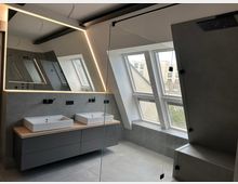 Modernes Badezimmer mit schräger Dachdecke, großem LED-beleuchtetem Spiegel und Doppelwaschtisch auf einem grauen Unterschrank. Rechts eine verglaste Dusche und mehrere Dachfenster, die Tageslicht und Blick auf umliegende Gebäude bieten.