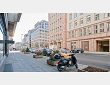 Straßenansicht in einer Stadt mit breitem Gehweg, parkenden Autos und einem Motorroller im Vordergrund. Die Straße ist umgeben von modernen mehrstöckigen Gebäuden mit großen Fenstern und rötlicher Fassade.