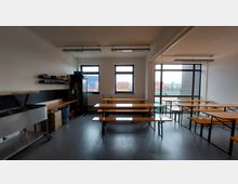 Ein einfacher Gemeinschaftsraum mit großen Fenstern, in dem mehrere Holzbänke und Tische aufgestellt sind. Links befindet sich eine Edelstahlspüle, Regale mit kleinen Gegenständen und ein Kühlschrank; durch die Fenster sind bunte Container und ein Hafengebiet zu sehen.