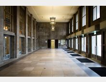 Großer Eingangsbereich mit hohen Decken, Boden aus hellen Steinfliesen und Wänden aus Betonplatten. Links befinden sich Glastüren und Fenster mit bunten Glaselementen, rechts eine Reihe von Glastüren mit Notausgangsschildern, die nach außen führen.