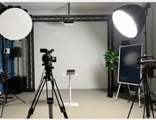 Professionell eingerichtetes Fotostudio mit einem weißen Hintergrund, zwei großen Softboxen als Lichtquellen, mehreren Kameras auf Stativen und einem kleinen weißen Podium in der Mitte. Im Hintergrund befinden sich eine grüne Pflanze und ein Monitor auf einem Ständer.