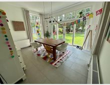 Esszimmer mit einem Holztisch, weißen Stühlen und einem bunten Teppich auf grauen Fliesen. Große Fenster und Glastüren bieten Blick auf einen grünen Garten, während bunte Geburtstagsdekorationen den Raum schmücken.