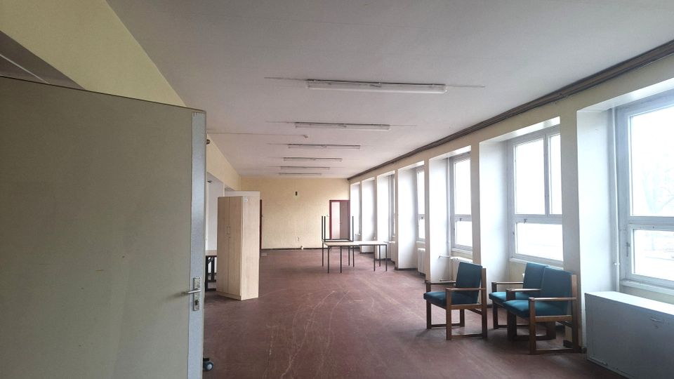 Langer, spärlich eingerichteter Innenraum mit braunem Boden, Decken-Leuchtstoffröhren und einer Reihe großer Fenster entlang der rechten Wand. Im Raum stehen wenige Möbel wie Stühle, Tische und ein Schrank; am Ende befindet sich eine Tür.