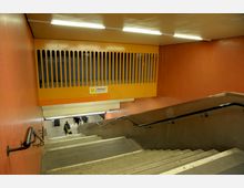 Treppenabgang zu einer U-Bahn-Station mit orange-roten Wänden und einer gelben Verkleidung mit vertikalen Lüftungsschlitzen. Im Hintergrund sind weitere Treppenstufen sowie zwei Personen in der Eingangshalle erkennbar.