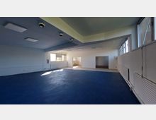 Großer, leerer Innenraum mit blauem Bodenbelag, weißen Wänden und rechteckigen Deckenleuchten. Der Raum verfügt über mehrere Fenster, Heizkörper entlang der Wände und eine offene Verbindung zu einem angrenzenden Bereich mit Fliesenboden.