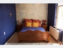 Schlafzimmer mit einem Holzbett im antiken Stil, farbigen Kissen und einer blauen Decke. Die Wände sind teils dunkelblau, teils mit verblasstem Putz versehen, flankiert von Nachttischen mit Lampen.