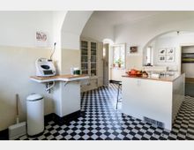 Helle Küche mit schwarzen und weißen Schachbrettfliesen, einer zentralen Kücheninsel mit Holzarbeitsplatte, mehreren weißen Schränken und einer Arbeitsfläche mit Fenster und Topfpflanzen. Im Hintergrund befinden sich gewölbte Durchgänge und ein Schrank mit Glastüren.