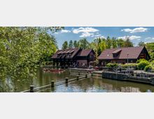 Ein Fluss mit mehreren roten Kanus im Vordergrund und zwei holzverkleideten Gebäuden mit roten Dachziegeln am Ufer, die als Bootsverleih dienen. Umgeben von Bäumen und Grünflächen unter klarem Himmel.