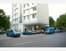 Mehrstöckiges Wohngebäude mit heller Fassade und einer offenen Eingangsüberdachung, umgeben von geparkten Autos auf einer gepflasterten Fläche. Im Hintergrund sind Bäume und eine Straße sichtbar.