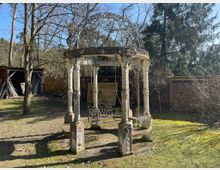 Steingartenpavillon mit einem verzierten Metallkuppel-Dach und gedrehten Steinsäulen, umgeben von einem Garten mit Bäumen und Holzlager an den Rändern. Im Hintergrund sind Holzschuppen und Zaunelemente zu sehen.