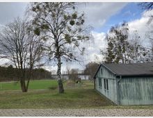 Freizeitbereich mit einer einfachen Holzhütte rechts im Bild, einem Fußballfeld mit Toren links und Bäumen im Hintergrund. Im Hintergrund ist ein Gewässer und ein bewölkter Himmel sichtbar.