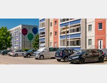 Parkplatz vor mehreren mehrstöckigen Wohnblocks mit Balkonen und farbigen Fassaden in Beige, Rot und Blau. Im Hintergrund ist ein Gebäude mit großer Wandmalerei aus drei Ballons zu sehen, daneben stehen Bäume unter blauem Himmel.