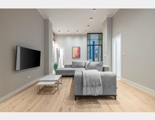 Modernes Wohnzimmer mit hellem Holzboden, grauen Wänden und hoher Decke mit Einbauspots, eingerichtet mit großem grauen Ecksofa und einem niedrigen Couchtisch. An der Wand hängt ein Flachbildfernseher; im Hintergrund führen Türen und eine verglaste Trennwand zu weiteren Räumen, ergänzt durch gerahmte Kunstwerke.