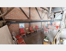 Ein industrieller Raum mit roten Backsteinwänden, Metallträgern und sichtbaren Rohren. Im Vordergrund stehen mehrere Stühle mit roten und beigen Polstern sowie runde Metalltische mit Glasplatten, die scheinbar für eine Versammlung oder Pause genutzt werden könnten.