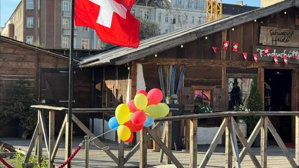 Holzhütte im Chalet-Stil auf einer erhöhten Holzterrasse mit Geländer, davor eine große Schweizer Flagge am Mast und ein Bündel bunter Luftballons. An der Fassade hängen kleine Schweizer Fähnchen und dekorative Skier; im Hintergrund stehen mehrstöckige Stadtgebäude.