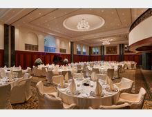 Großer, eleganter Bankettsaal mit runden, weiß eingedeckten Tischen, umgeben von gepolsterten Stühlen. Die Decke ist mit Kronleuchtern und versteckten Lichtern verziert, während die Wände Holztäfelungen, große Rundbögen und dekorative Elemente aufweisen; im Hintergrund steht ein schwarzer Flügel.