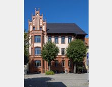 Historisches Gebäude mit hell verputzter Fassade und Zierelementen aus rotem Backstein, verziert durch einen turmartigen Giebel. Vor dem Gebäude stehen mehrere Bäume, und der gepflasterte Vorplatz ist teilweise im Schatten.