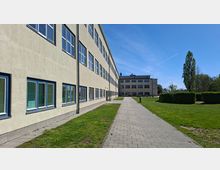 Langer, mehrstöckiger Gebäudekomplex mit heller Fassade und vielen rechteckigen Fenstern entlang eines gepflasterten Weges. Rechts liegen Rasenflächen mit geschnittenen Hecken und Bäumen, im Hintergrund steht ein weiteres Gebäude unter blauem Himmel.