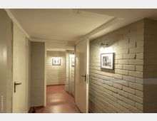 Flur in einem Innenbereich mit hell gestrichenen Backsteinwänden, rotem Bodenbelag und weißen Türen. An den Wänden hängen Fotos in Rahmen, die von kleinen Wandleuchten beleuchtet werden.