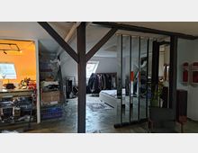 Dachgeschosswohnung mit sichtbaren Holzbalken und einem offenen Grundriss. Im Vordergrund befindet sich ein Bereich mit Spiegelpaneelen, dahinter ein Schlafbereich mit Bett, Kleiderstange und einem kleinen Teppich; rechts ist eine Sitzecke und links eine Küche mit Neonlicht zu sehen.