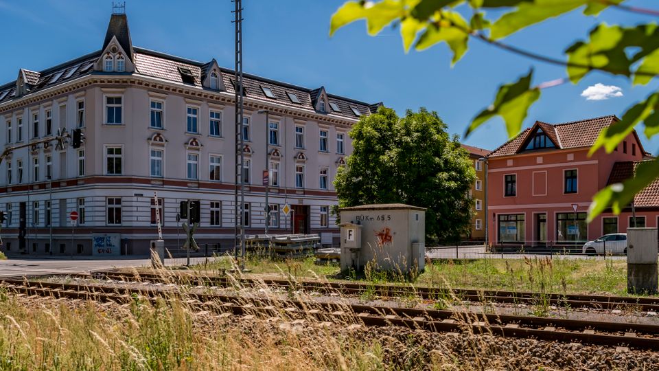 Straßenszene an einem Bahnübergang mit mehreren Gleisen im Vordergrund, hohem Gras und Signalanlagen. Dahinter stehen ein großes, historisches mehrstöckiges Eckgebäude mit dekorierter Fassade und Dachgauben sowie kleinere Häuser mit roten Ziegeldächern unter blauem Himmel.