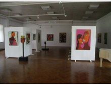 Ein heller Ausstellungsraum mit weißen Wänden und Holzfußboden, in dem Gemälde und Skulpturen ausgestellt sind. Die Kunstwerke sind auf Stellwänden und Sockeln präsentiert, während von der Decke Schienenlampen herabhängen, die den Raum beleuchten.