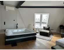 Modernes Dachgeschosszimmer mit freistehender Whirlpool-Badewanne in dunkler Marmorverkleidung, hellem Holzfußboden und sichtbaren schwarzen Deckenbalken. Neben einem Doppelfenster stehen eine graue Liege auf Teppich, ein kleiner Beistelltisch und eine Laterne mit Kerzen.
