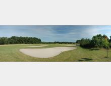 Ein Golfplatz mit kurz gemähtem Rasen, mehreren Sandbunkern und einer Baumreihe im Hintergrund. Der Himmel ist leicht bewölkt, und die Landschaft wirkt offen und weitläufig.