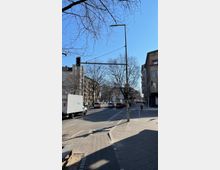 Straßenszene in einer Stadt mit einer Ampel und mehreren geparkten sowie fahrenden Autos. Auf beiden Seiten der Straße befinden sich mehrstöckige Gebäude, im Hintergrund sind kahle Bäume sichtbar, und der Himmel ist klar und blau.