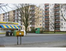 Straßenansicht mit einem gelben Briefkasten im Vordergrund, Recyclingcontainern und einer kleinen Ladenfront mit gelbem und blauem Vordach links. Im Hintergrund sind mehrstöckige Wohngebäude mit Balkonen sowie einige kahle Bäume zu sehen.