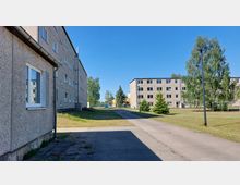 Mehrstöckige Wohngebäude aus Betonplatten in einer gepflegten Grünanlage mit Rasenflächen, kleinen Bäumen und Gehwegen, umgeben von vereinzelten Straßenlaternen; im Hintergrund sind weitere Gebäude und ein Zaun sichtbar.