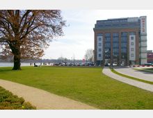 Große Rasenfläche mit einem Kiesweg und einem großen Baum im Vordergrund, im Hintergrund ein mehrstöckiges modernes Bürogebäude mit der Aufschrift „Oracle“ und ein Fluss mit Spaziergängern entlang des Ufers.