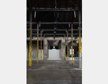 Interior of a large empty industrial warehouse with exposed steel roof trusses, hanging light fixtures, and rows of tall yellow support columns. Weathered brick walls and a wide open concrete floor lead toward a white partition wall at the far end.