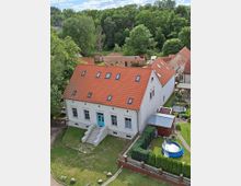 Historisches Gebäude mit weißer Fassade und rotem Satteldach, umgeben von einem Garten mit Rasen, Hecken und einem Pool. Im Hintergrund dichter Wald und eine Backsteinmauer.