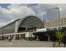 Bahnhof Alexanderplatz in Berlin mit einer modernen Glasfassade und gewölbtem Dach. Im Hintergrund sind das Hochhaus des "Park Inn"-Hotels sowie umliegende Gebäude sichtbar.