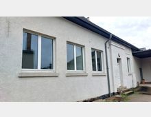 Weiße, einstöckige Gebäudefassade mit mehreren rechteckigen Fenstern und einer Eingangstür. Das Gebäude hat ein flaches Satteldach, eine Außenbeleuchtung über der Tür und einen gepflasterten Vorbereich.