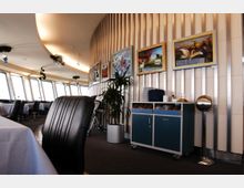 Innenansicht eines modernen Restaurants oder Cafés mit großen Fensterfronten, die schräg geneigte Scheiben zeigen. An einer gewölbten Holzverkleidung mit vertikalen Streben hängen mehrere Bilder, und ein mobiler Schrank steht daneben; im Vordergrund befindet sich ein gepolsterter Stuhl.