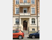 Fassade eines mehrstöckigen Wohngebäudes im Altbaustil mit gelblich-braunem Mauerwerk, weißen Zierleisten und einem säulenbetonten Eingangsportal mit brauner Holztür. Vor dem Gebäude stehen zwei geparkte Autos, ein orangenes und ein blaues, auf einer gepflasterten Straße.