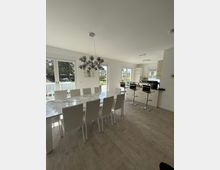 Modernes Esszimmer mit offener Küche. Ein großer Esstisch mit weißem Marmor-Effekt und passenden weißen Stühlen steht im Vordergrund, dahinter eine Küchenzeile mit Barhockern und großen Fenstern, die viel Tageslicht einlassen.