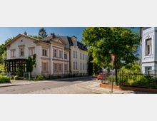 Straßenszene in einem ruhigen Wohnviertel mit historischen Gebäuden im Altbaustil, verziert mit Stuckelementen und Pastellfarben. Im Vordergrund steht ein großer Baum neben einer gepflasterten Straße, eine Vorfahrtsregelung ist durch ein Verkehrsschild markiert.