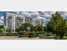 Mehrstöckiges Wohngebäude im Hintergrund mit weißen Fassaden und Balkonen, davor eine Grünanlage mit Bäumen und Sträuchern sowie ein Parkplatz mit geparkten Autos; rechts ein Straßenschild mit der Aufschrift „Platz der Befreiung“.