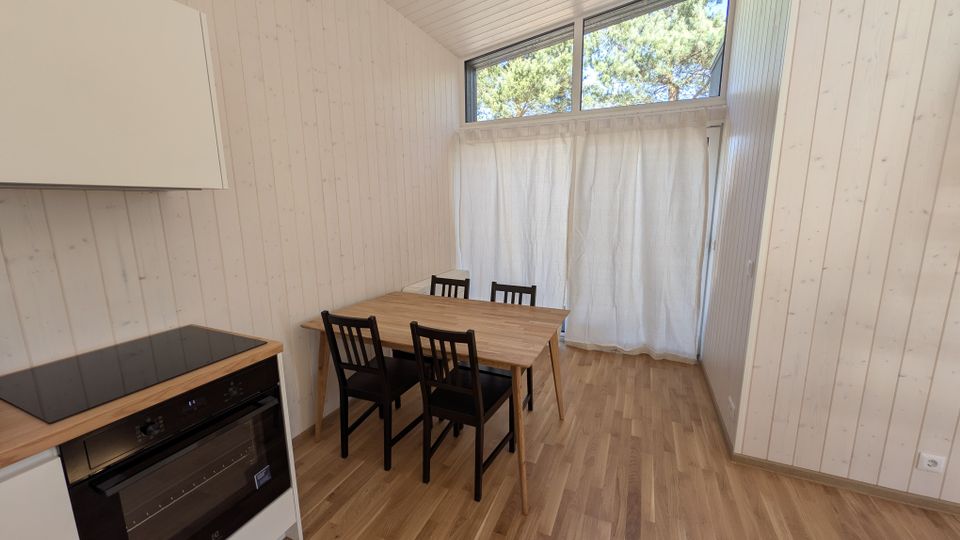 Heller Essbereich in einem schmalen Raum mit weißen Holzvertäfelungen, Holzfußboden und einem kleinen Tisch mit vier Stühlen neben einer Küchenzeile. Am Ende des Raums befindet sich eine bodennahe Fensterfront mit Vorhängen und einem hohen Oberlichtfenster mit Blick auf Bäume.